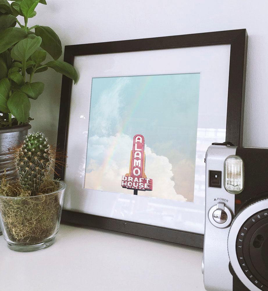 Framed Alamo Drafthouse Austin Art print by Betsy Crum showing vintage theater sign against blue sky displayed with plants and camera