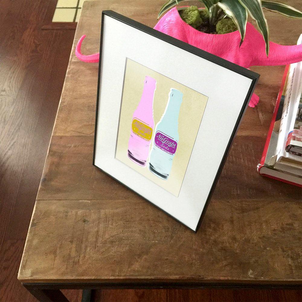Framed pop art museum print of NuGrape Soda Bottles in Warhol and Lichtenstein style on wooden table.