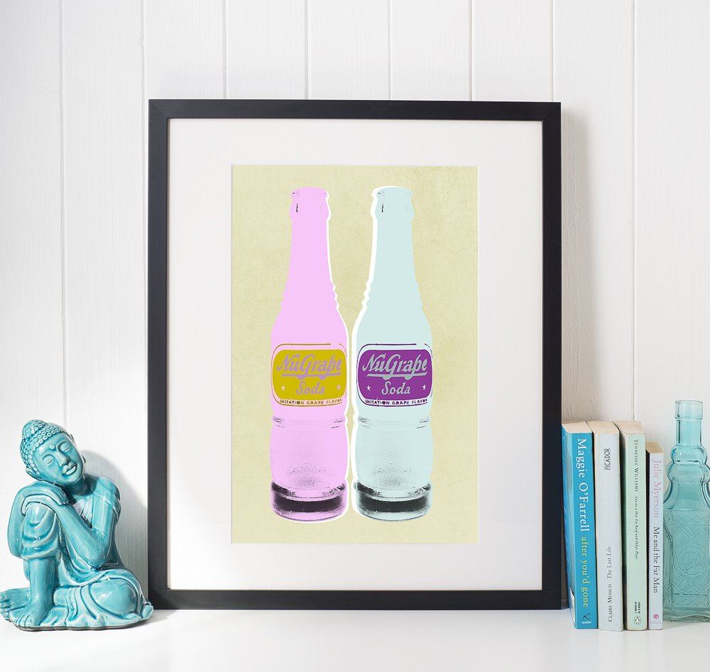 Framed pop art print of two vintage NuGrape soda bottles in pastel colors displayed on a wall next to books and a turquoise Buddha sculpture