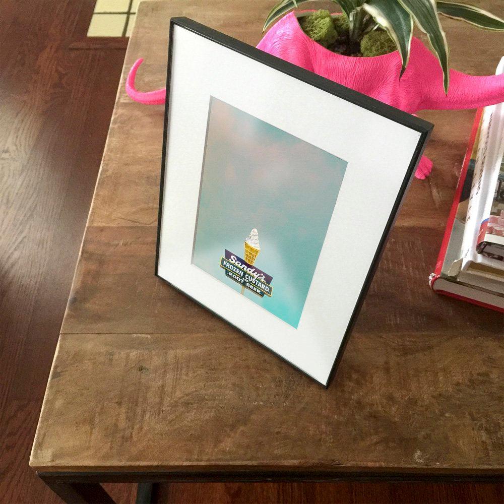 Framed museum print of Sandys Ice Cream Austin Texas sign on wooden table next to pink planter and books