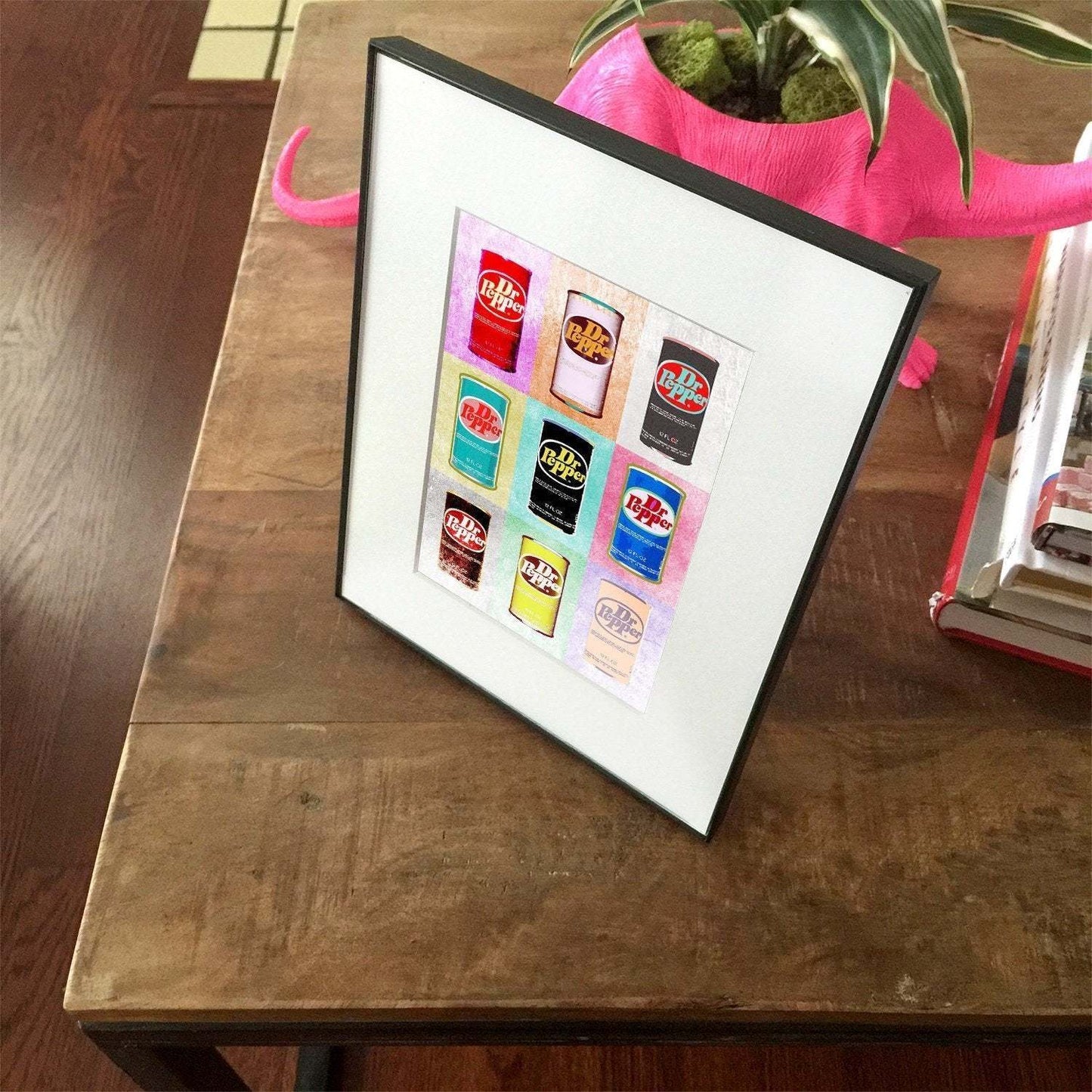 Framed Dr. Pepper Warhol style print with colorful soda cans on a wooden table next to a pink planter and books