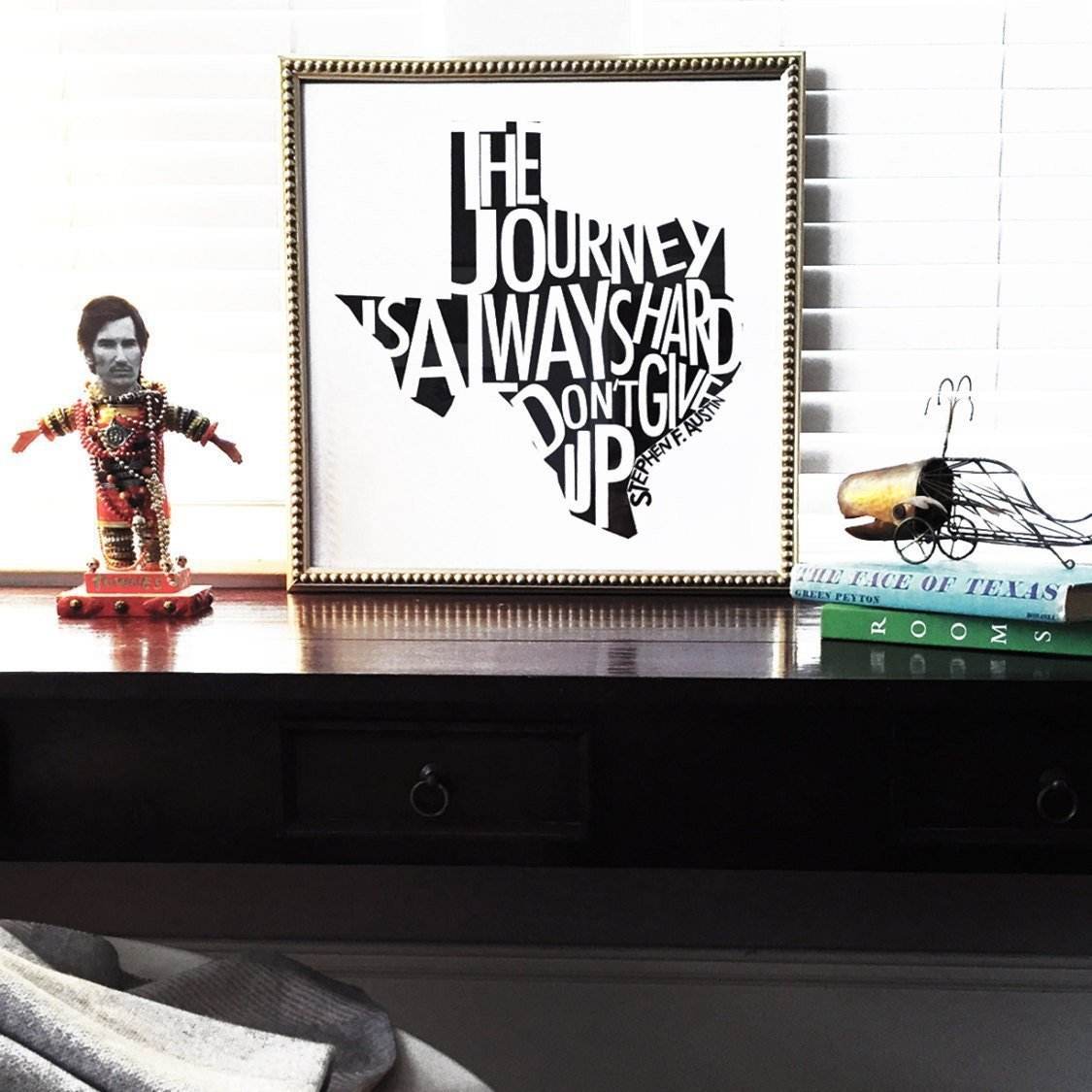 Framed Stephen F. Austin Quote print reading "The Journey is Always Hard, Don't Give Up" in Texas-shaped typography on desk with books and decorative figurine