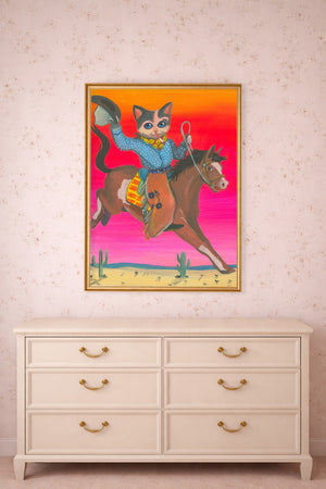 Smoking Cat Art Print in a gold frame on a beige to pink wall below a bedroom dresser.  This Cowboy Cat is on a horse and is considered a western wall art.  Prints up to  24x36" of this Western Cat Art Print by Caya Crum.