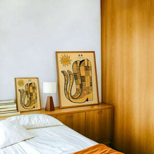 Roach Stompers cowboy boot art print by Caya Crum — framed in a wood-accent room