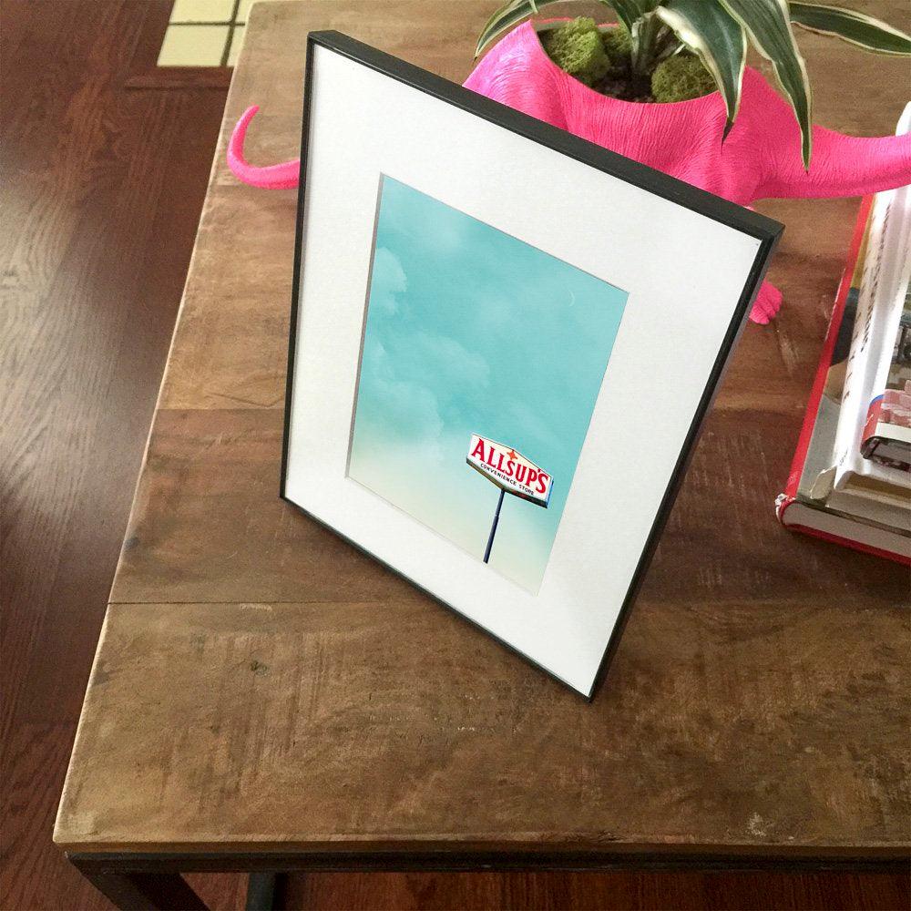 New Mexico Art. Framed Allsups Art print featuring a blue sky with the Allsups sign on a wooden table