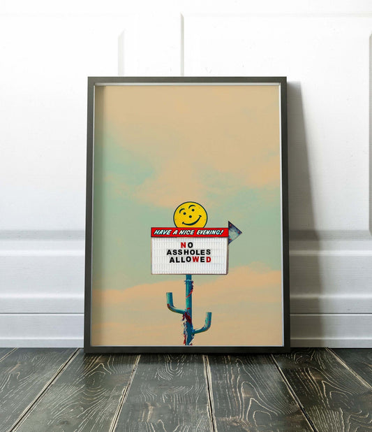  No Assholes Allowed Print Framed vintage-style roadside sign print with smiley face on muted sky background leaning against white wall on wooden floor