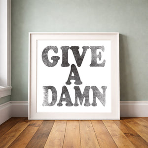 Give a Damn black on white print by Betsy Crum framed upright against a grey-green wall