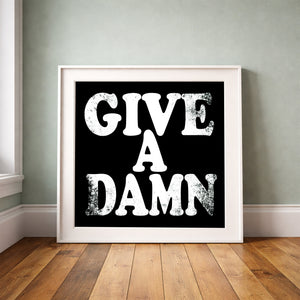 Give a Damn white on black print by Betsy Crum framed upright against a grey-green wall
