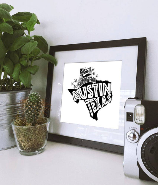 Greetings From Austin Texas black and white art print in black frame with plants and camera on white surface