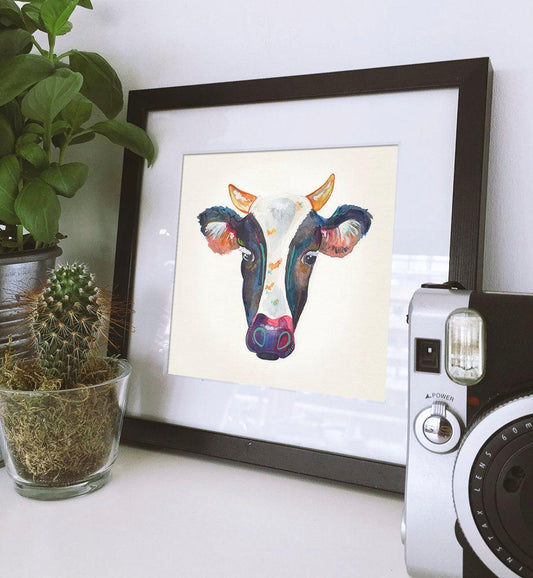 Framed colorful cow head art print displayed on a table with potted plants and instant camera nearby