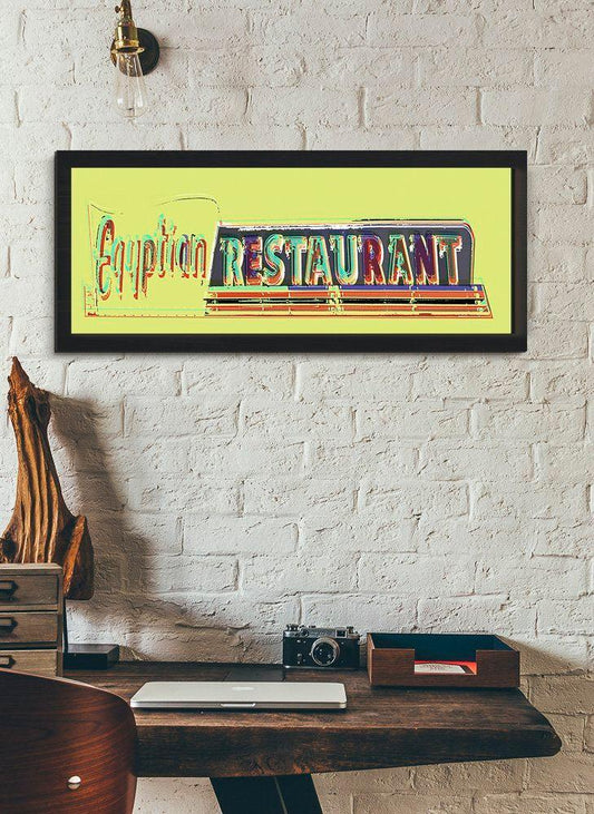 Framed museum print of a colorful neon sign reading Egyptian Restaurant on a yellow background hanging on a white brick wall above a wooden desk.