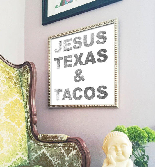 Framed museum print with distressed text reading "Jesus Texas & Tacos" displayed on a wall next to a vintage green patterned chair and a small Buddha statue with plants.