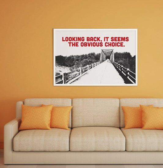 Museum print titled "Looking Back, It Seems the Obvious Choice" featuring a bridge illustration with red text above, framed and displayed above a beige sofa.