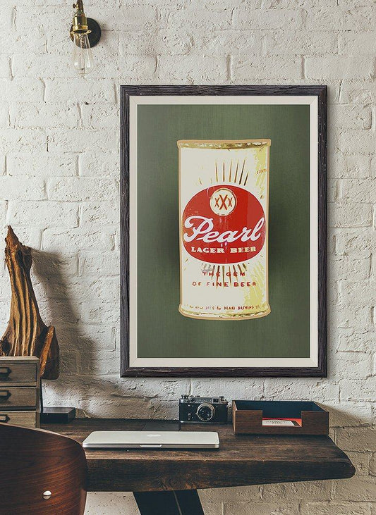 Framed art print of Pearl Beer Can by Betsy Crum on green background displayed on brick wall above wooden desk