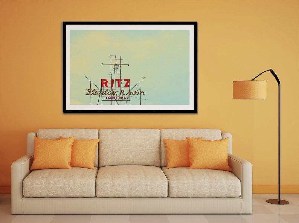Ritz Starlite Room art print by Betsy Crum in black frame above beige sofa with orange pillows and lamp