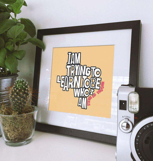 Stevie Ray Vaughan art print with "I Am Trying To Learn To Be Who I Am" text in black frame on beige background, displayed on a white surface with potted plants and vintage camera.