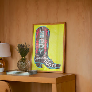 Leddy Boot Yellow cowboy boot art print by Betsy Crum — framed in a wood-accent room